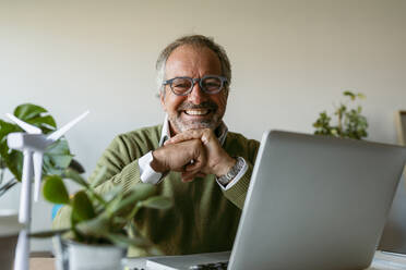 Lächelnder Mann mit Brille, der einen Laptop benutzt, während er zu Hause am Tisch sitzt - VABF03623