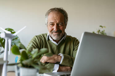 Älterer Mann mit verschränkten Armen, der einen Laptop benutzt, während er zu Hause am Tisch sitzt - VABF03624