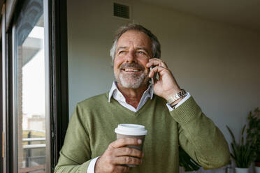 Man with coffee cup talking on mobile phone while standing at home - VABF03629