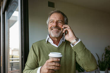 Mann mit Kaffeetasse, der zu Hause steht und mit seinem Handy telefoniert - VABF03629