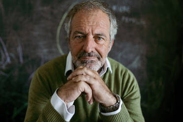 Mature man with hand on chin sitting against blackboard at home - VABF03635