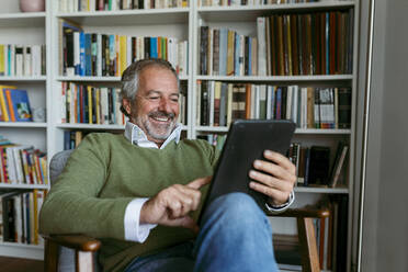 Lächelnder Mann, der ein digitales Tablet benutzt, während er zu Hause am Bücherregal sitzt - VABF03640