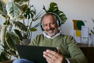 Ältere Männer tragen Kopfhörer, während sie zu Hause ein digitales Tablet benutzen - VABF03643