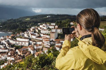 Rückansicht einer Frau, die im Urlaub mit ihrem Smartphone ein Dorf fotografiert - DMGF00202