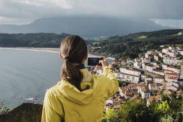 Rückansicht einer Frau, die im Urlaub mit dem Handy ein Dorf fotografiert - DMGF00203