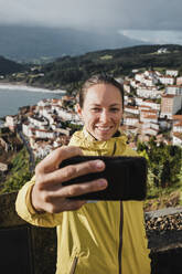 Lächelnde Frau nimmt Selfie mit Dorf durch Smartphone während der Ferien - DMGF00205