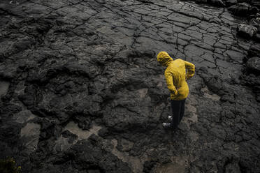 Drohnenaufnahme einer Frau, die einen Regenmantel trägt, während sie in der Regenzeit auf felsigem Boden steht - DMGF00210