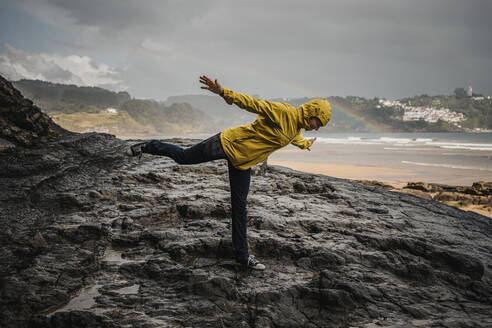Playful woman wearing raincoat while standing on rocky ground during rainy season - DMGF00211
