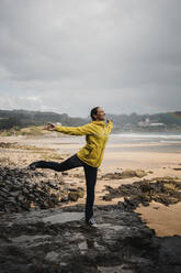 Playful woman in raincoat standing on on one leg at beach during rainy season - DMGF00215