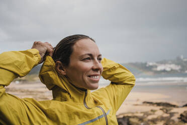 Smiling woman wearing raincoat at beach during rainy season - DMGF00216