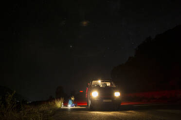 Weiblicher Camper sitzt vor einem Minivan mitten auf einer Landstraße in der Nacht - MAMF01366