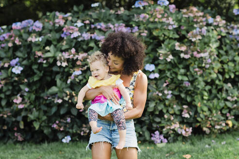 Mittlere erwachsene Frau, die ein kleines Mädchen trägt, während sie vor einer blühenden Pflanze im Park steht - NMSF00422