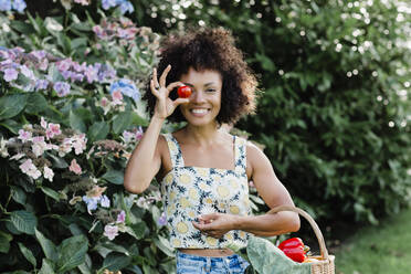 Lächelnde erwachsene Frau, die eine Tomate hält, während sie an einer blühenden Pflanze im Park steht - NMSF00427
