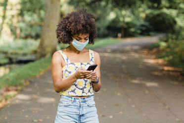 Mittlere erwachsene Frau mit Gesichtsmaske, die ein Mobiltelefon benutzt, während sie auf der Straße im Park steht - NMSF00428