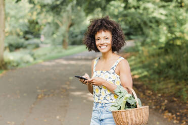 Lächelnde Frau, die einen Gemüsekorb hält, während sie ein Mobiltelefon im Park benutzt - NMSF00430