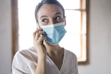 Businesswoman wearing face mask talking on mobile phone while standing at office - MOEF03480