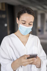 Geschäftsfrau mit Gesichtsmaske, die ein Mobiltelefon benutzt, während sie im Büro steht - MOEF03483