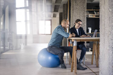 Geschäftsfrau, die einen Laptop benutzt, während ein Mann auf einem Fitnessball im Büro sitzt - MOEF03484