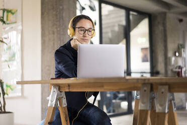 Geschäftsfrau mit Kopfhörern, die einen Laptop benutzt, während sie am Tisch im Büro sitzt - MOEF03498