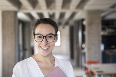 Smiling businesswoman wearing eyeglasses holding digital tablet while standing at office - MOEF03517