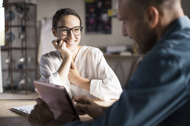 Lächelnde Frau, die einen Mann ansieht, der ein digitales Tablet benutzt, während er im Büro sitzt - MOEF03530