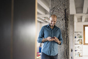 Lächelnder Geschäftsmann, der ein Mobiltelefon benutzt, während er an einer Säule im Büro steht - MOEF03535