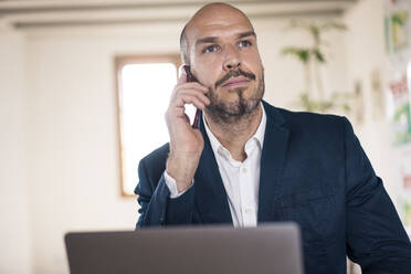 Geschäftsmann, der mit seinem Handy telefoniert, während er im Büro am Laptop arbeitet - MOEF03544