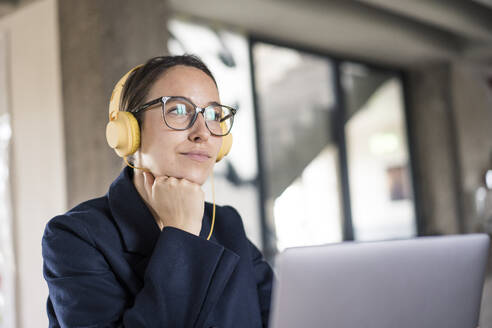 Geschäftsfrau mit Kopf in den Händen und Kopfhörern, die einen Laptop benutzt, während sie im Büro sitzt - MOEF03549