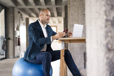 Geschäftsmann, der ein Mobiltelefon und einen Laptop benutzt, während er auf einem Fitnessball im Büro sitzt - MOEF03553