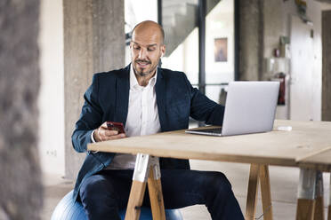 Mann im Anzug, der Laptop und Handy benutzt, während er im Büro sitzt - MOEF03554