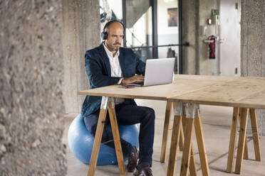 Geschäftsmann mit Kopfhörern, der einen Laptop benutzt, während er auf einem Fitnessball im Büro sitzt - MOEF03558