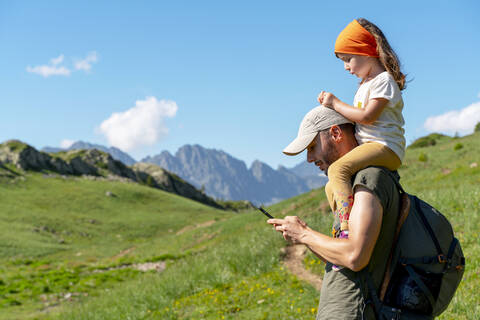Father using smart phone while daughter sitting on his shoulder during sunny day stock photo