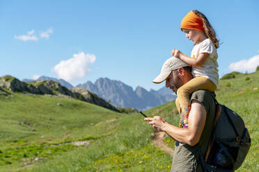 Father using smart phone while daughter sitting on his shoulder during sunny day - GEMF04294