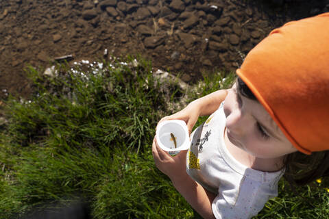 Cute little girl holding disposable cup with fish inside it standing on lakeshore stock photo