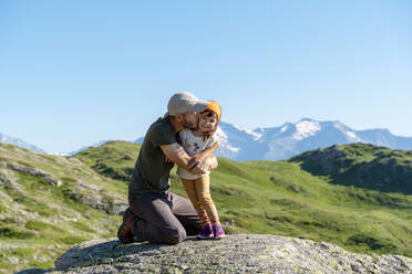 Vater küsst seine Tochter auf einem Felsen kniend an einem sonnigen Tag - GEMF04306