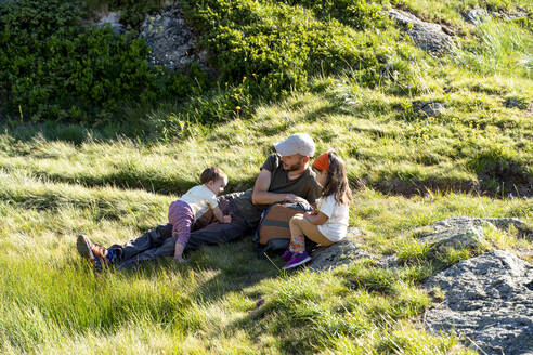 Vater verbringt seine Freizeit mit seinen Töchtern im Gras sitzend an einem sonnigen Tag - GEMF04311