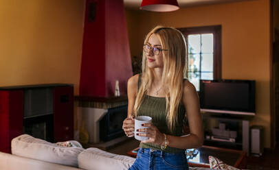 Nachdenkliches Teenager-Mädchen, das eine Kaffeetasse hält, während es zu Hause wegschaut - MGOF04593