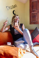 Fröhliche Frau hält Handy und Brille, während sie zu Hause auf dem Sofa sitzt - MGOF04607