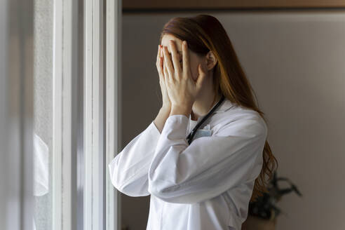 Emotionally stressed young female doctor with head in hands standing by window - AFVF07385