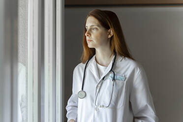 Nachdenkliche junge Ärztin, die durch das Fenster schaut, während sie in ihrem Büro zu Hause steht - AFVF07386