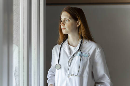 Thoughtful young female doctor looking through window while standing at home office - AFVF07386
