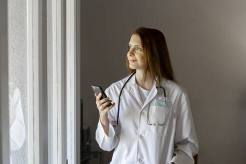 Smiling young female doctor looking through window while holding smart phone - AFVF07388
