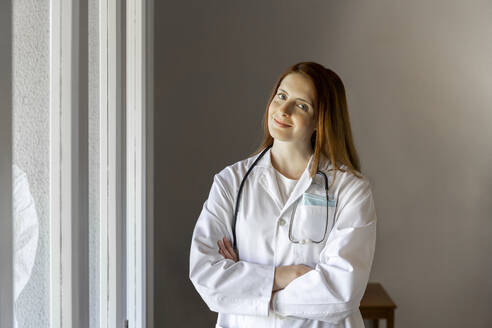 Confident young female doctor standing with arms crossed at home office - AFVF07389