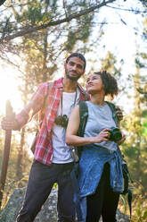 Happy young couple hiking with camera and binoculars in sunny woods - CAIF29824