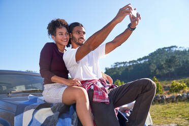 Glückliches junges Paar macht ein Selfie auf der Motorhaube eines Autos am sonnigen Straßenrand - CAIF29828