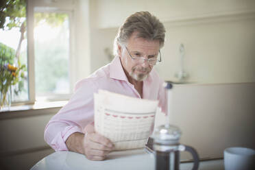 Älterer Mann liest Zeitung am Laptop am morgendlichen Küchentisch - CAIF29837