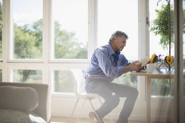 Senior man reading book at dining table by sunny window - CAIF29841