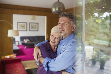 Glückliches älteres Paar umarmt sich am sonnigen Wohnzimmerfenster - CAIF29843