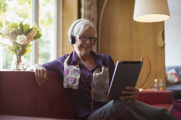 Smiling senior woman with headphones using digital tablet on sofa - CAIF29847
