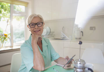 Porträt einer selbstbewussten älteren Frau, die ein Smartphone in der Morgenküche benutzt - CAIF29852